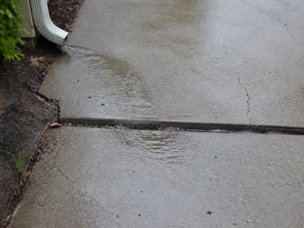 Rainwater moving from a downspout across a low concrete driveway joint.